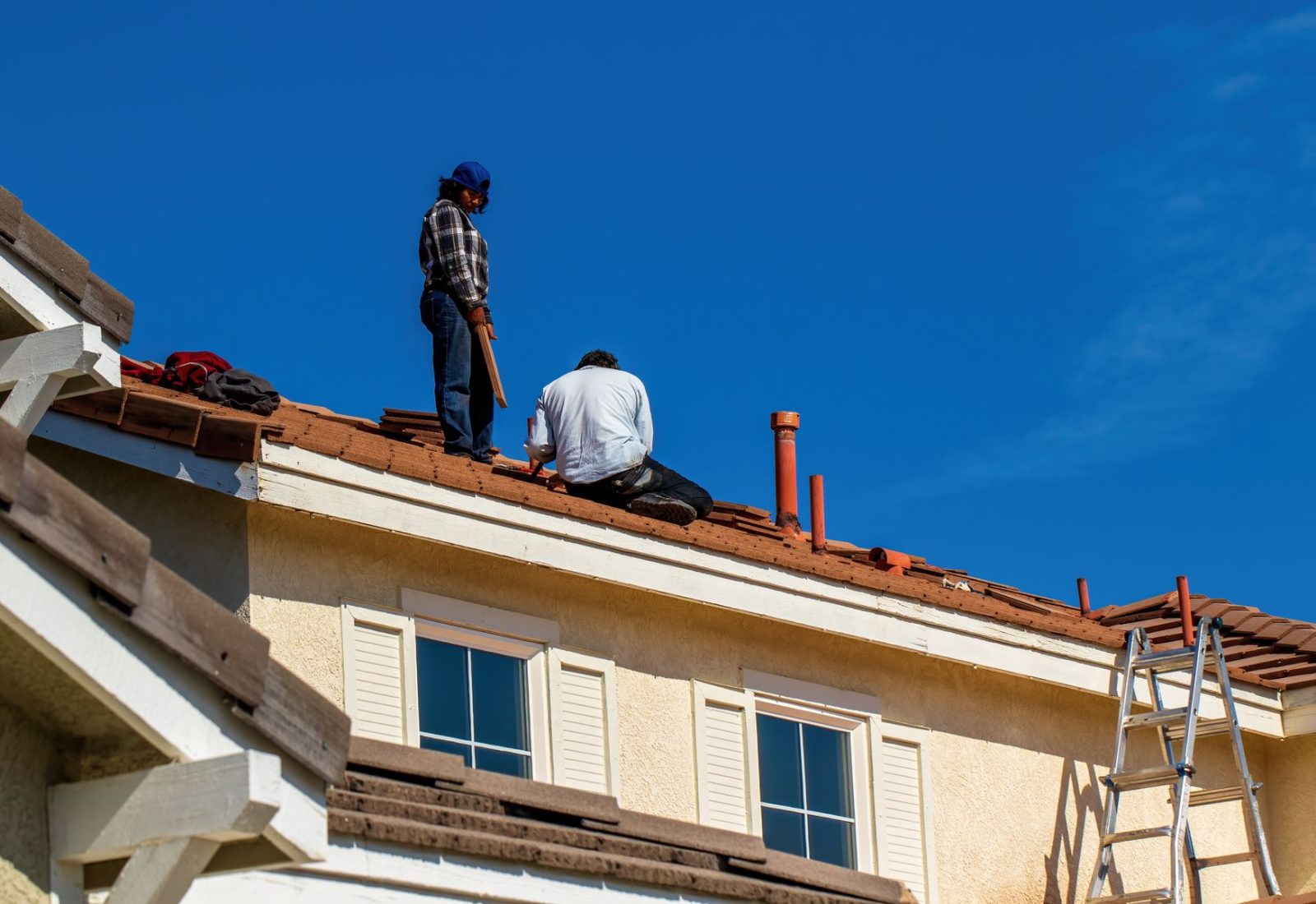 Zwei Dachdecker sitzen bei der Arbeit auf einem Dach unter blauem Himmel