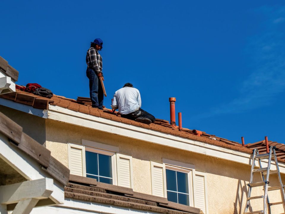 Zwei Dachdecker sitzen bei der Arbeit auf einem Dach unter blauem Himmel