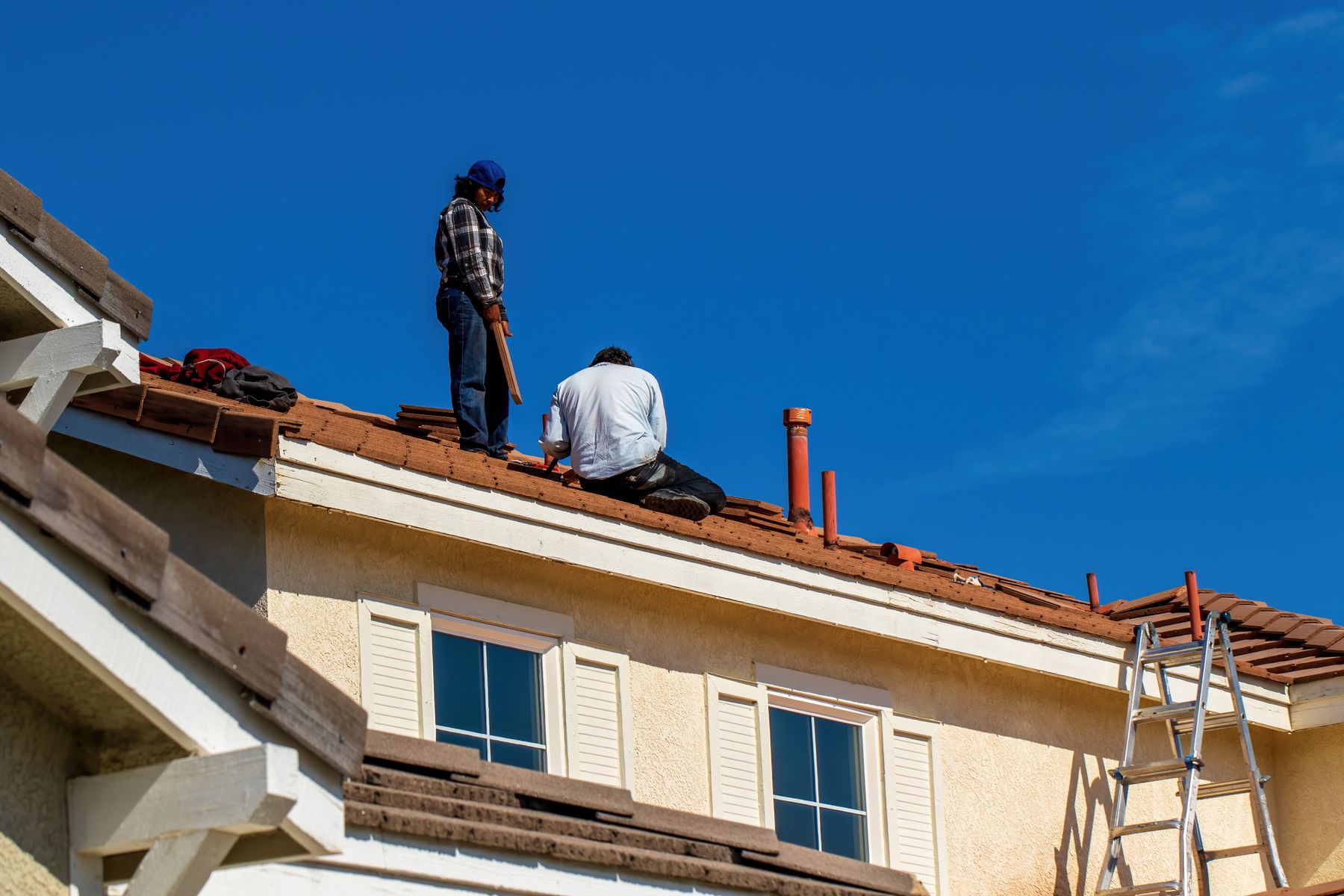 Zwei Dachdecker sitzen bei der Arbeit auf einem Dach unter blauem Himmel